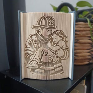 May include: A folded book art sculpture featuring a firefighter wearing a helmet with the number 23, carrying a small child in their arms. The design is created by precisely folding the pages of a book to form a three-dimensional image.