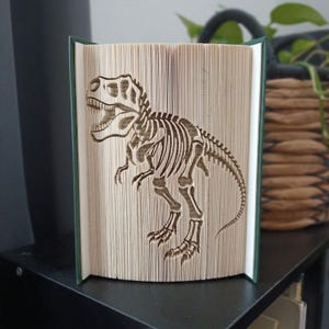 May include: A book art sculpture featuring a folded page design that creates the silhouette of a Tyrannosaurus Rex skeleton. The pages are precisely creased to form the dinosaur's skull, ribcage, spine, and tail, set within a dark green book cover.