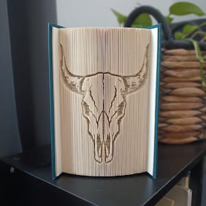 May include: A book art sculpture featuring a folded page design that creates the silhouette of a bull skull. The book has a teal cover and the pages are cream-coloured, with the intricate skull shape formed by precise folds in the paper edges.