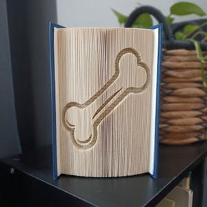 Può includere: Una scultura in libro piegato raffigurante la forma di un osso di cane, creata piegando con precisione le singole pagine di un libro con copertina rigida blu. Le pagine sono color crema e formano una chiara silhouette di osso centrata.