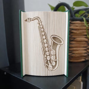Op de afbeelding: Een boek met gevouwen pagina's die een driedimensionale saxofoonvorm creëren. Het boek heeft een donkergroene kaft en de pagina's zijn wit met zichtbare tekst. Het saxofoonontwerp is gecentreerd op de open pagina's van het boek.