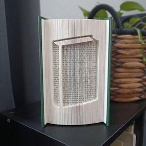 Puede incluir: Una escultura de libro plegado que presenta un diseño central recortado de un marco rectangular con una barra superior. Las páginas están dobladas con precisión para formar esta figura geométrica, con el texto impreso original visible.