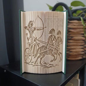 Può includere: Una scultura realizzata con un libro piegato che mostra la silhouette dettagliata di una figura dell'antico Egitto su un carro trainato da due cavalli. La figura impugna arco e freccia, con le pagine del libro piegate per formare l'immagine.