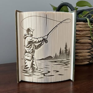 Op de afbeelding: Een boeksculptuur met ingewikkeld gevouwen pagina's die het silhouet vormen van een persoon die aan het vliegvissen is in het water. Het ontwerp toont een figuur met een pet en vest, die een hengel met een gebogen lijn vasthoudt.