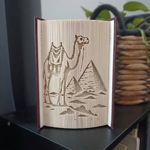 Puede incluir: Una escultura de libro con páginas dobladas de forma intrincada que forman la silueta de un camello junto a dos pirámides. El libro tiene una cubierta de color granate oscuro y las páginas son de color crema, mostrando texto.