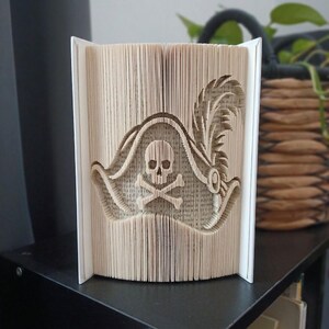 Könnte beinhalten: Eine Buchfaltkunst-Skulptur, die einen dreidimensionalen Piratenhut mit einem Totenkopf- und Knochenemblem in der Mitte darstellt. Die Seiten sind präzise gefaltet, um die Form zu erzeugen, mit einem Federdetail auf der rechten Seite.