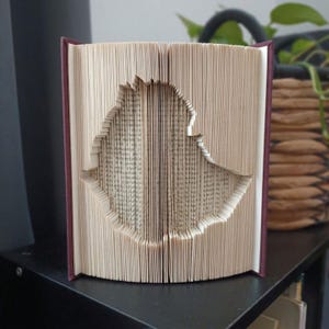 Peut inclure: Un livre aux pages pliées formant la silhouette tridimensionnelle d'un oiseau. Le livre possède une couverture bordeaux foncé et les pages sont de couleur crème avec du texte imprimé en noir. Les pages sont pliées pour former l'oiseau.