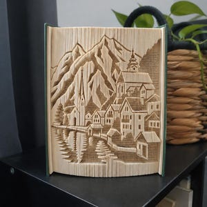 Könnte beinhalten: Eine Buchskulptur mit gefalteten Seiten, die eine dreidimensionale Szene eines Bergdorfes bilden. Das Design umfasst einen hohen Kirchturm, mehrere Häuser und gezackte Berggipfel im Hintergrund, die alle aus Papier geformt sind.