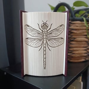 Op de afbeelding: Een boeksculptuur met een libelleontwerp, gemaakt door de pagina's van een hardcover boek te vouwen. Het boek heeft een donkerrode kaft en de witte pagina's zijn nauwkeurig gevouwen om het ingewikkelde, symmetrische silhouet van een libelle te tonen.