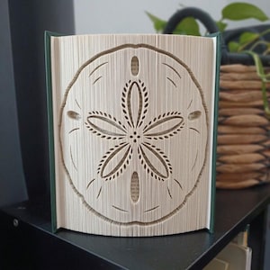 Pode incluir: Uma escultura de arte em livro dobrado com um design de bolacha-da-praia, criado dobrando com precisão as páginas de um livro. A escultura tem uma capa verde-escura e exibe o padrão simétrico intrincado de cinco pétalas.