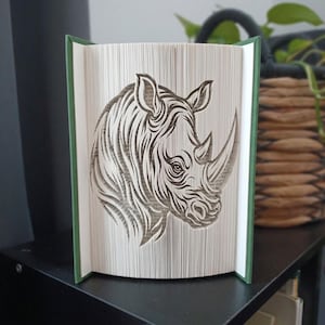 Op de afbeelding: Een boeksculptuur met een gevouwen paginadesign dat het profiel van een neushoornkop vormt. Het boek heeft een donkergroene kaft en de pagina's zijn nauwkeurig gevouwen om de gedetailleerde contouren, oren en hoorn van het dier te tonen.