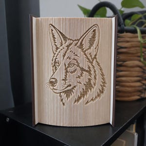 Op de afbeelding: Een boeksculptuur met een gevouwen paginadesign dat een gedetailleerd, driedimensionaal portret van een wolvenkop vormt. De pagina's zijn crèmekleurig met donkerbruine kaften aan de zijkanten, wat nauwkeurige papier-vouwtechnieken laat zien.