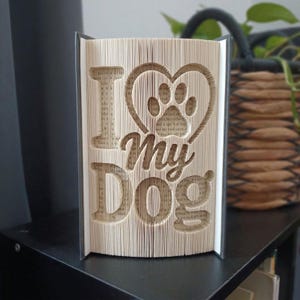 Puede incluir: Una escultura de libro plegado con el texto I heart my dog, donde el corazón tiene forma de huella de perro. Las páginas están dobladas con precisión para crear las letras tridimensionales y el diseño gráfico de los bordes del papel.