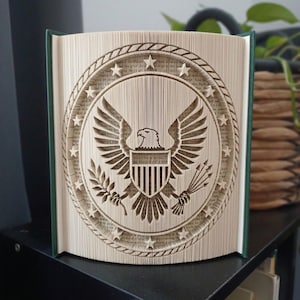 May include: A folded book art sculpture featuring the Great Seal of the United States. The pages are precisely creased to form a central eagle with outstretched wings, a shield on its chest, olive branches, arrows, and a surrounding ring of stars.