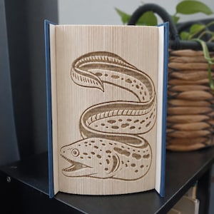 Könnte beinhalten: Eine Buchskulptur mit einer Faltkunst-Gestaltung, die eine gefleckte Muräne darstellt. Die Seiten sind präzise gefaltet, um die Form des Aals zu bilden, der in einem Schlangenmuster gewunden ist. Das Buch hat dunkelblaue Einbände an den Seiten.