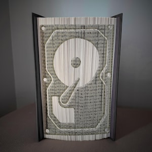 Pode incluir: Escultura de arte de livro única com um design de disco rígido. As páginas são meticulosamente dobradas para criar uma imagem tridimensional de um disco rígido. O livro está em pé, mostrando os intrincados detalhes do design.
