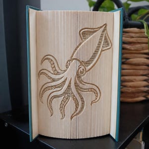 Könnte beinhalten: Eine Buchkunstskulptur mit einem Tintenfisch-Motiv, das durch Falten der Seiten eines Hardcover-Buches entsteht. Die Seiten sind fächerförmig geöffnet, um die komplexe Form des Tintenfischs zu zeigen, wobei die blaugrünen Buchdeckel sichtbar sind.