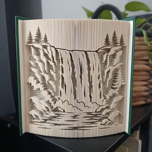 Puede incluir: Una escultura de libro con páginas dobladas intrincadamente que forman una escena de cascada tridimensional. El diseño incluye formaciones rocosas en capas, un efecto de agua en cascada y árboles de hoja perenne a ambos lados, hechos de papel.