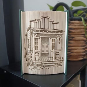 Può includere: Una scultura in libro piegato che raffigura un dettagliato edificio in stile saloon western con porte a battente, un portico, una ruota di carro e una lanterna. Le pagine sono piegate con precisione per formare la struttura, tra copertine verde scuro.