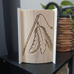 Può includere: Una scultura realizzata con un libro piegato che presenta un design ritagliato di un gioco per gatti a bacchetta con due piume pendenti. Le pagine sono piegate con precisione per creare l'immagine, mostrando il testo sui bordi interni.