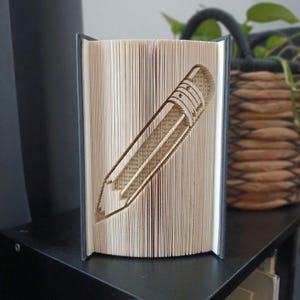 Op de afbeelding: Een boeksculptuur met pagina's die zijn gevouwen om de duidelijke vorm van een potlood te creëren. Het boek staat rechtop met de donkergrijze kaften aan de zijkanten zichtbaar, wat de ingewikkelde papier-vouwtechniek voor het potloodontwerp toont.