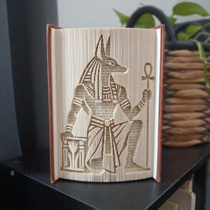 May include: A book sculpture featuring a folded page art design of the Egyptian deity Anubis. The figure is depicted with a jackal head, wearing traditional headdress and garments, holding an ankh staff in one hand. The pages are cream colored.