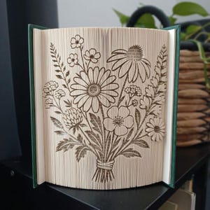 May include: A folded book art sculpture featuring a detailed bouquet of wildflowers. The pages are precisely creased and cut to form the silhouettes of daisies, clover, and various stems, creating a three-dimensional floral arrangement.