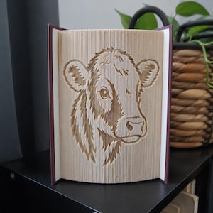 Könnte beinhalten: Eine Buchfaltkunst-Skulptur, die ein detailliertes, dreidimensionales Porträt eines Kuhkopfes zeigt. Die Buchseiten sind präzise gefaltet, um das Bild des Tieres zu erzeugen, wobei die dunklen Buchdeckel an der linken und rechten Seite sichtbar sind.