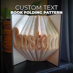 May include: A book with pages folded to spell "Custom" in a 3D design. The book is open, revealing the folded text, with the words "Custom Text Book Folding Pattern" above the book.