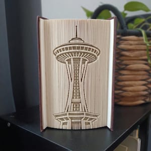 May include: A book sculpture featuring the Space Needle in Seattle, created by precisely folding the pages of a hardcover book. The cream-colored paper pages are manipulated to form the iconic tower silhouette, showcasing intricate structural details.
