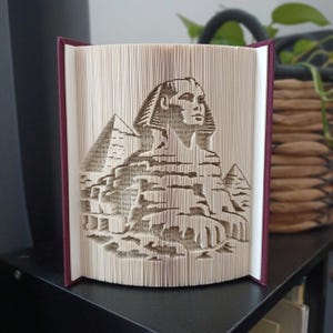 May include: A folded book art sculpture featuring the Great Sphinx of Giza and pyramids. The pages are precisely creased and folded to create a three-dimensional relief image of the Egyptian monument, set within a dark maroon hardback book cover.