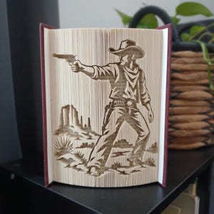 Könnte beinhalten: Eine gefaltete Buchkunst-Skulptur, die einen Cowboy mit Hut und Halstuch zeigt, der eine Pistole hält und in einer Wüstenlandschaft mit Kakteen und Felsformationen steht. Die Seiten sind präzise gefaltet, um die Silhouette der Figur zu bilden.