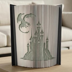 May include: A book sculpture with a dragon and castle design. The pages are cut and folded to create a 3D image of a dragon in flight above a castle with multiple towers. The book has a black cover and is displayed on a wooden table.