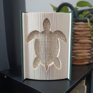 May include: A folded book art sculpture featuring a sea turtle silhouette created by precisely folding the individual pages of a hardcover book. The book has a dark green cover, and the interior pages display visible printed text throughout.