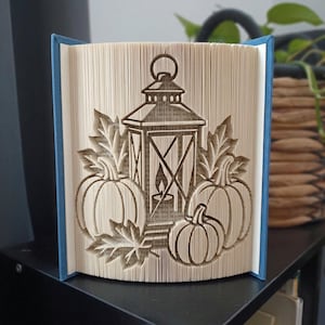 Könnte beinhalten: Eine Buchfaltkunst-Skulptur mit einem zentralen Laternenmotiv, flankiert von drei Kürbissen und mehreren Blättern. Die Seiten sind präzise gefaltet, um das dreidimensionale Bild zu erzeugen, wobei blaue Buchdeckel an den Seiten sichtbar sind.