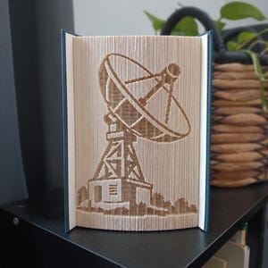 Könnte beinhalten: Eine Buchfaltkunst-Skulptur mit einem detaillierten Satellitenschüssel-Design, das durch präzises Falten der einzelnen Seiten eines Hardcover-Buches entsteht. Die cremefarbenen Seitenkanten bilden die Form der Schüssel und ihre Stützstruktur.