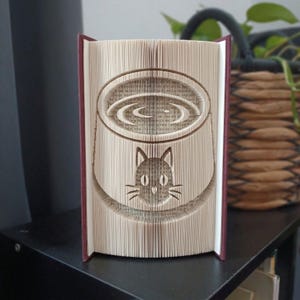 Peut inclure: Une sculpture en livre plié représentant un visage de chat sous une forme circulaire. Les pages sont pliées avec précision pour former l'image, avec les couvertures de livre bordeaux visibles sur les côtés gauche et droit de la structure.