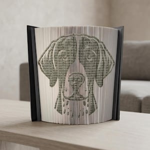 Op de afbeelding: Een decoratief boek met een hondenhoofd dat in de pagina's is gesneden. De kop van de hond staat centraal op het boek, met de pagina's uitgespreid om de afbeelding te creëren. Het boek heeft een zwarte rug en staat op een lichtgekleurd oppervlak.