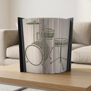 May include: A decorative book sculpture featuring a drum set design. The artwork is created by fanning the pages of a book to reveal the image of a drum kit. The book is encased in a black frame and sits on a wooden table.