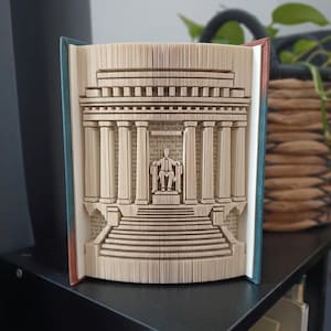 May include: A book sculpture featuring folded pages that form the shape of a neoclassical building with columns, a wide staircase, and a central seated figure. The book covers are visible on the sides, showing a teal and reddish-brown colour scheme.