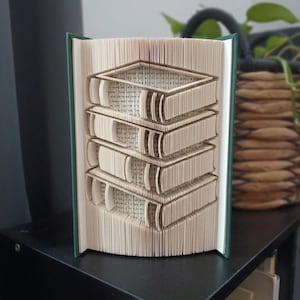 May include: A book sculpture featuring folded pages that form the shape of a stack of four books. The book has a dark green cover and the pages are cream-colored with visible black printed text throughout the folded paper structure.
