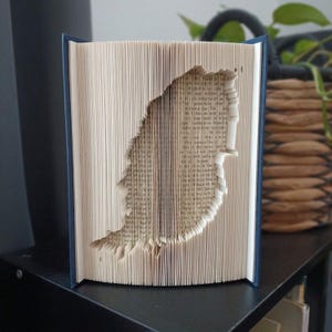 May include: A book with folded pages creating a three-dimensional cutout shape of the island of Ireland. The book has a dark blue cover and the interior pages are cream-colored with black printed text visible throughout the folded paper structure.