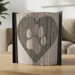 Peut inclure: Un livre décoratif dont les pages sont pliées pour créer un cœur avec une empreinte de patte découpée. Les pages du livre sont gris clair, et le cœur et l'empreinte de patte sont gris plus foncé. Le livre a une tranche noire et est présenté sur une surface en bois.