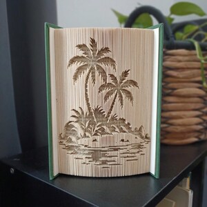 以下が含まれることがあります： 2本のヤシの木と低い植生が描かれた熱帯の島をモチーフにした、折り本アートの彫刻。ページは正確に折り畳まれており、木々と島の立体的なシルエットを作り出しています。緑色のブックカバーの中に収められています。