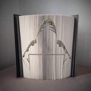 Pode incluir: Uma peça de arte de livro única com um design recortado de uma pessoa usando um capuz. As páginas são meticulosamente dobradas e cortadas para criar a silhueta. O livro tem uma capa cinza e o design recortado é em tons de verde e cinza.