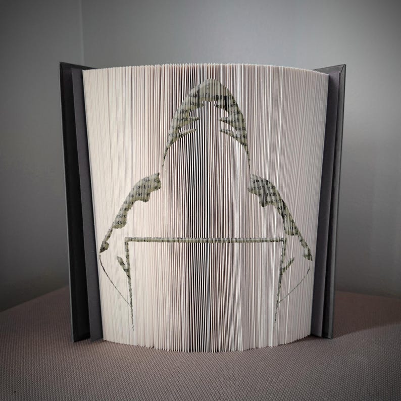 Peut inclure: Une &oelig;uvre d'art unique en forme de livre repr&eacute;sentant un personnage portant un sweat &agrave; capuche. Les pages sont pli&eacute;es et coup&eacute;es avec pr&eacute;cision pour cr&eacute;er la silhouette. Le livre a une couverture grise et le motif d&eacute;coup&eacute; est dans des tons de vert et de gris.