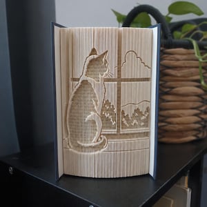 Könnte beinhalten: Eine Buchskulptur mit gefalteten Seiten, die eine dreidimensionale Silhouette einer Katze auf einem Fensterbrett bilden. Das Design zeigt die Katze, die durch einen Fensterrahmen auf eine Landschaft mit Wolken und Bäumen in der Ferne blickt.