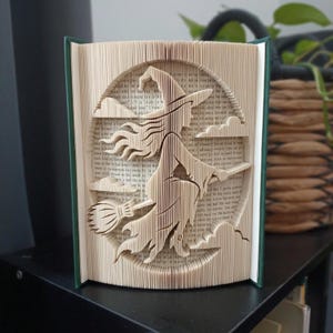 May include: A book sculpture featuring folded pages that form the silhouette of a witch flying on a broomstick. The design includes clouds and a circular background, all created by precisely folding the individual pages of a hardcover book.