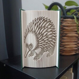 May include: A book sculpture featuring a folded page art design of an echidna. The pages are precisely creased to form the silhouette of the spiny animal, with the dark green book covers visible on the left and right sides of the open volume.