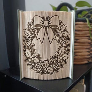 Pode incluir: Uma escultura de livro dobrado apresentando um design de guirlanda detalhado com um laço proeminente no topo, pinhas e folhas de azevinho. As páginas são dobradas com precisão para criar a imagem tridimensional na borda frontal do livro.